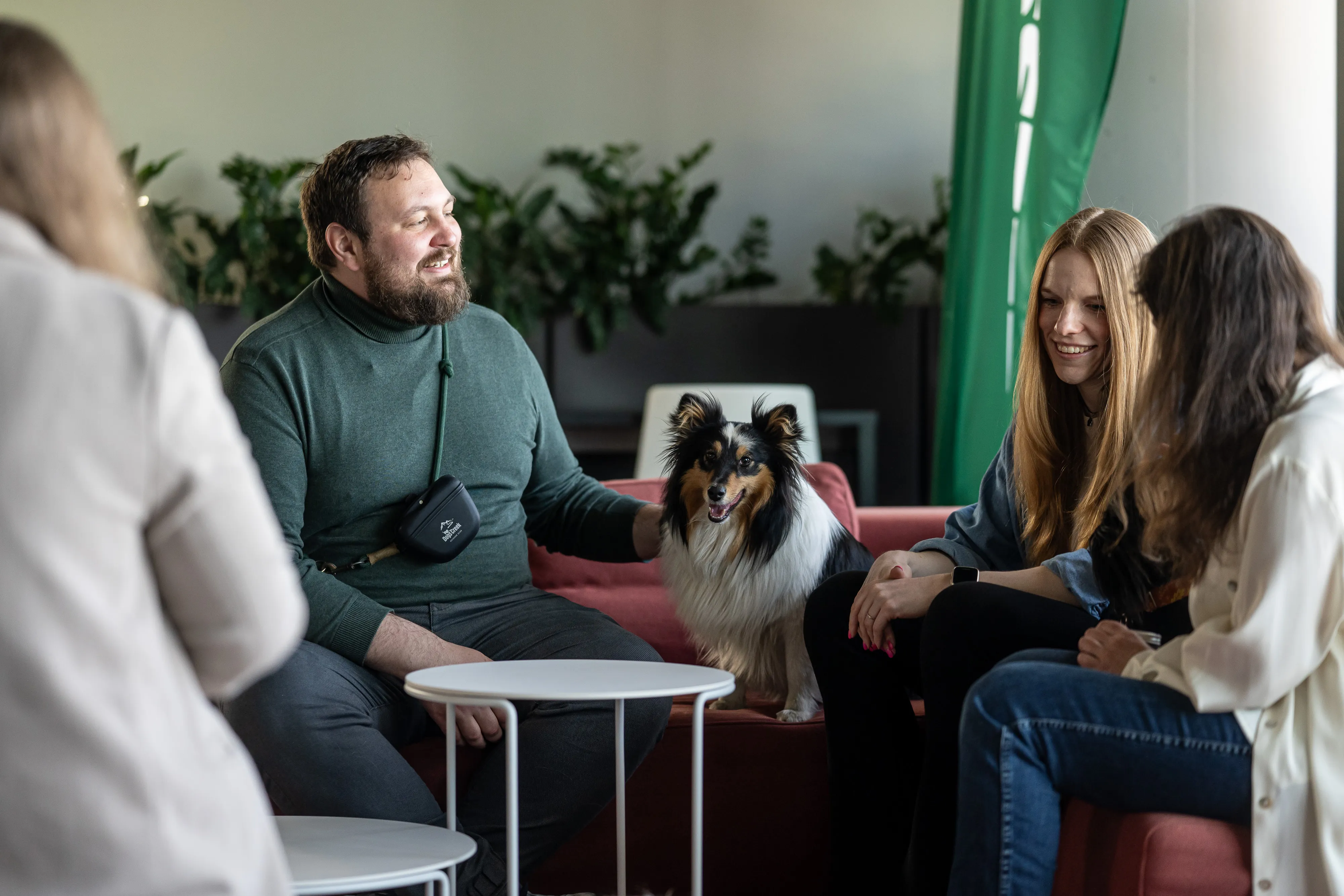 A group of coworkers hanging out on a couch with a friendly dog, chatting and smiling in a relaxed office vibe.