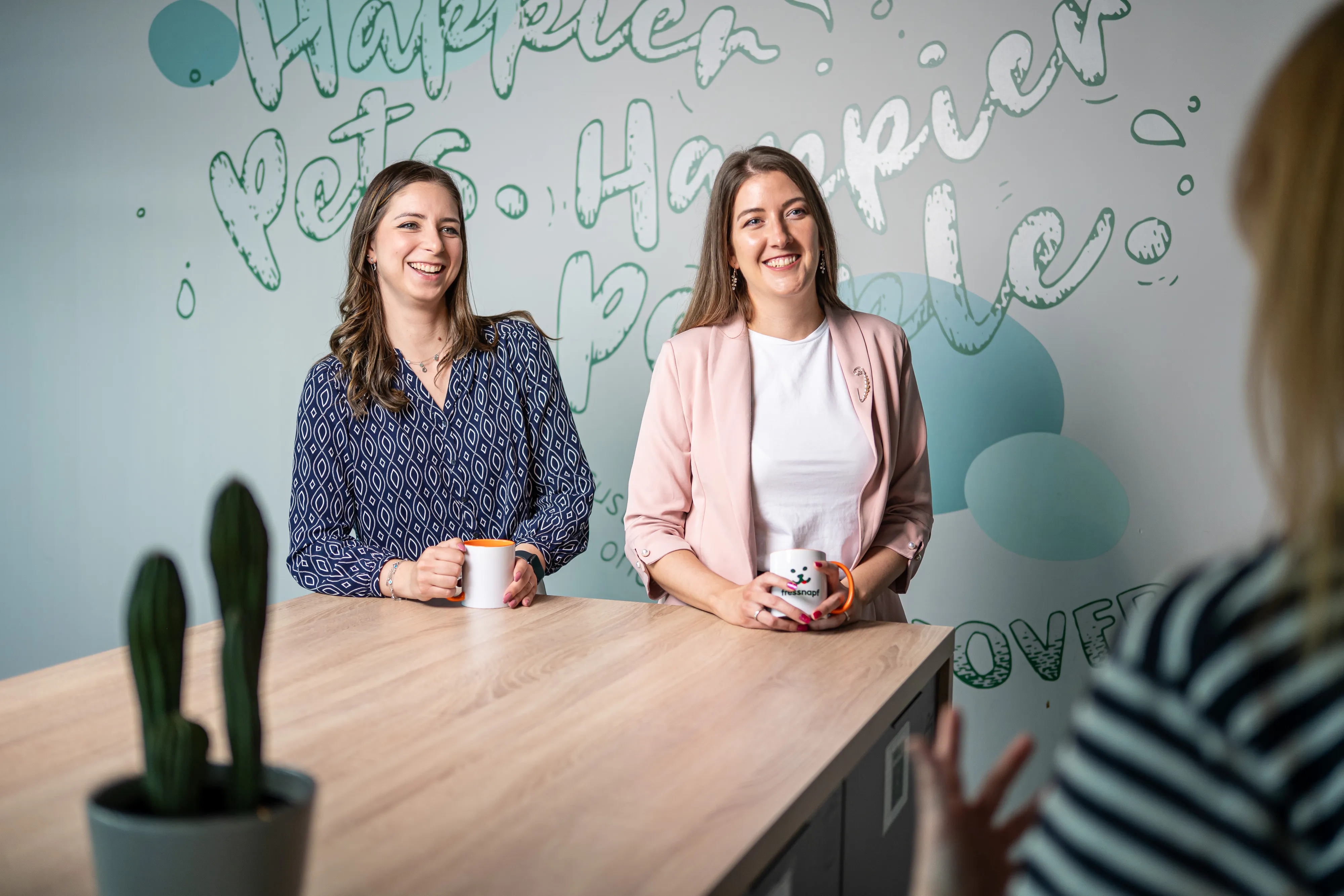 Two smiling women chat over coffee mugs in a casual Fressnap International office, with friendly wall art behind them.