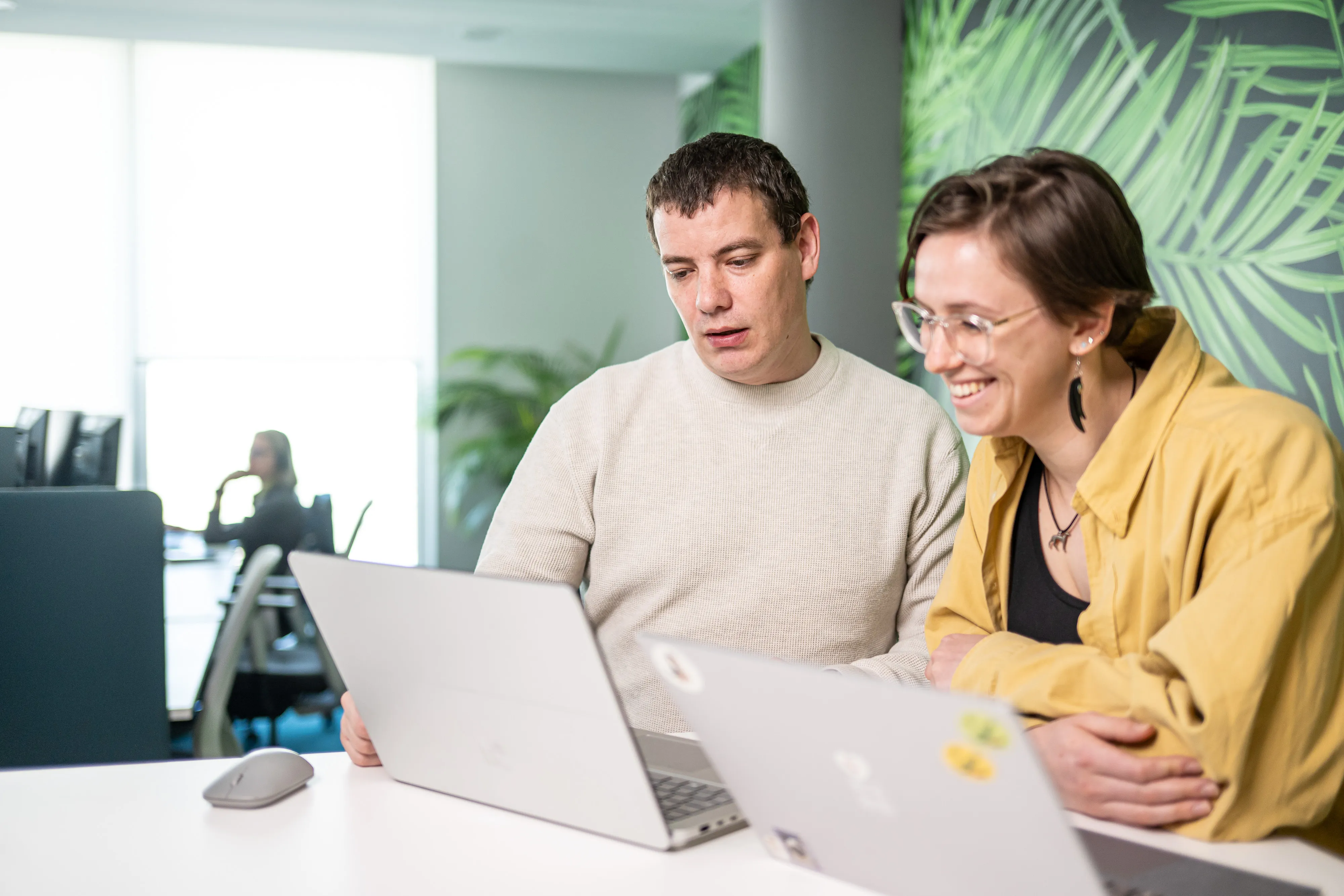 Two coworkers chatting and laughing while working together on laptops in a bright, casual office space.