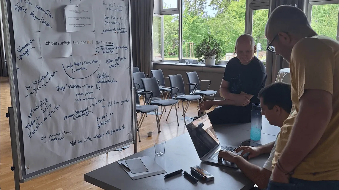 Drei IT-Kollegen arbeiten gemeinsam am Laptop, neben einem Whiteboard mit Brainstorming-Ideen im modernen Seminarraum.
