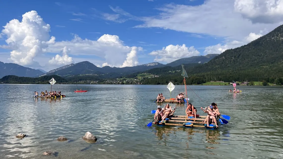 Gepardec-Team beim Floßbau auf einem See, umgeben von Bergen und blauem Himmel – erlebe Teamspirit hautnah.