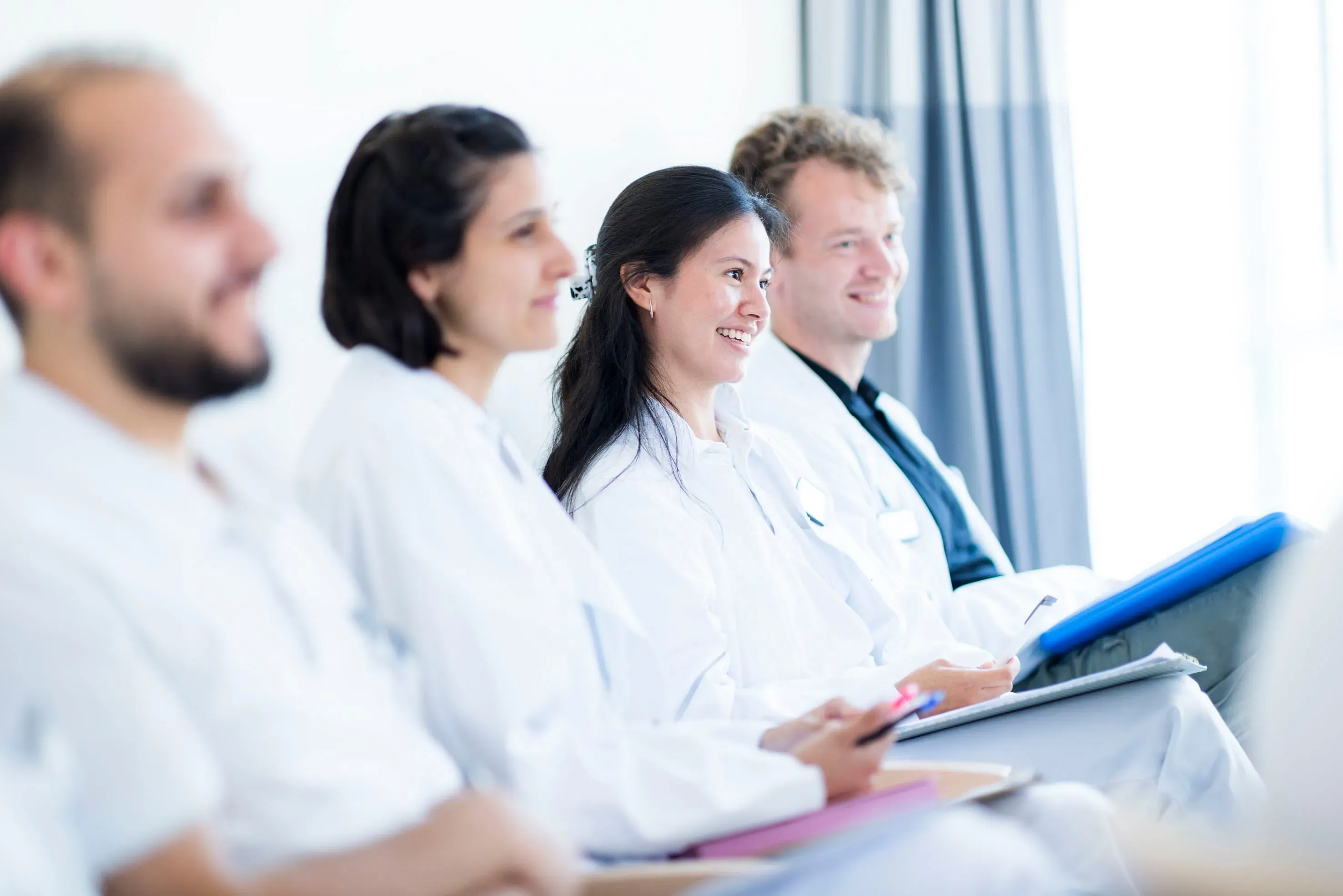 Vier junge Menschen in weißer Berufskleidung lächeln und hören aufmerksam zu.