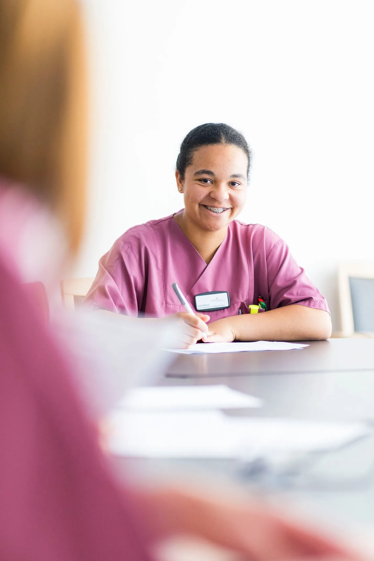 Junge Pflegekraft in rosa Kasack lächelt und schreibt bei einer Besprechung