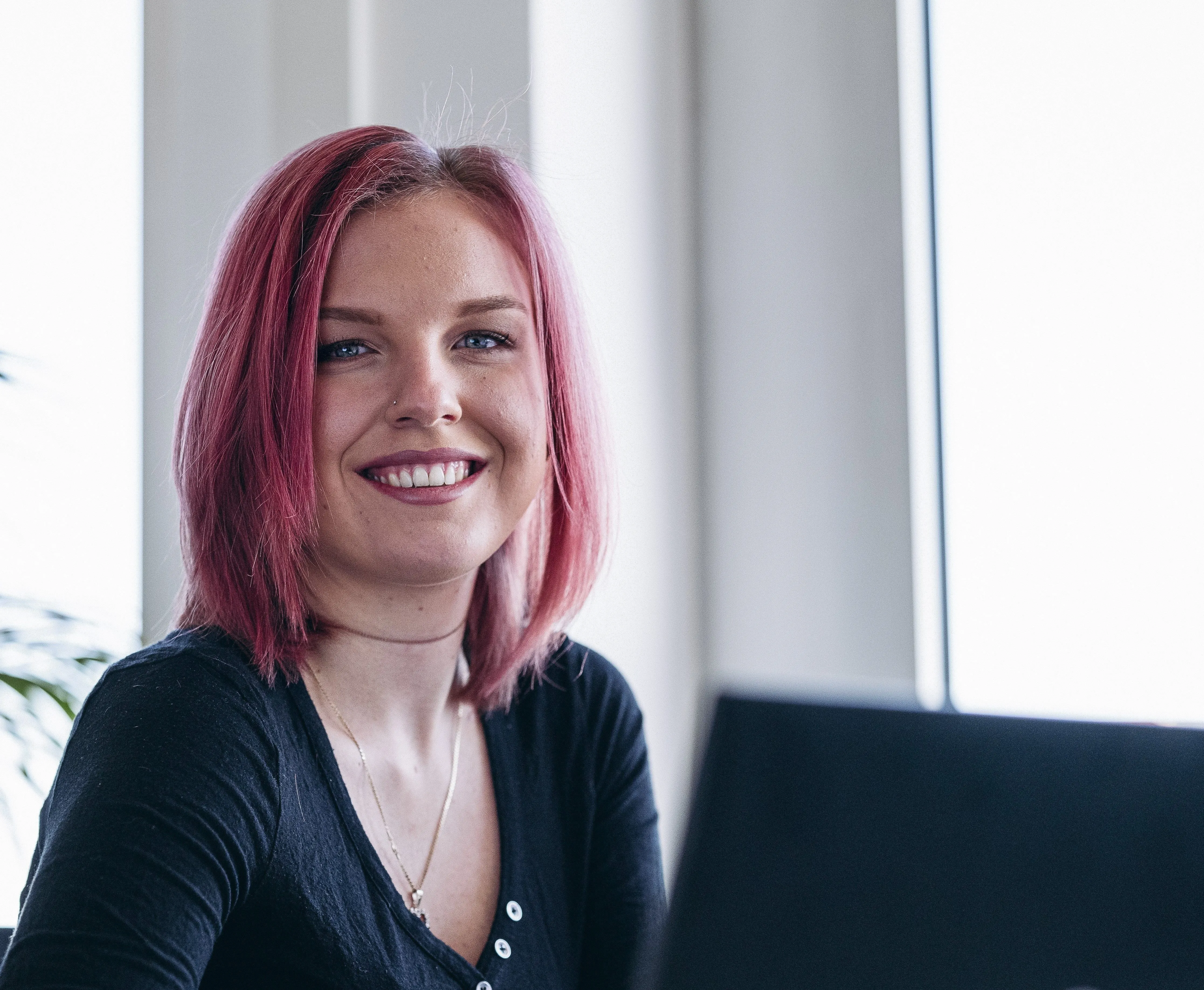 Eine lächelnde Frau mit pinken Haaren sitzt in einem Büro, vor einem Laptop.