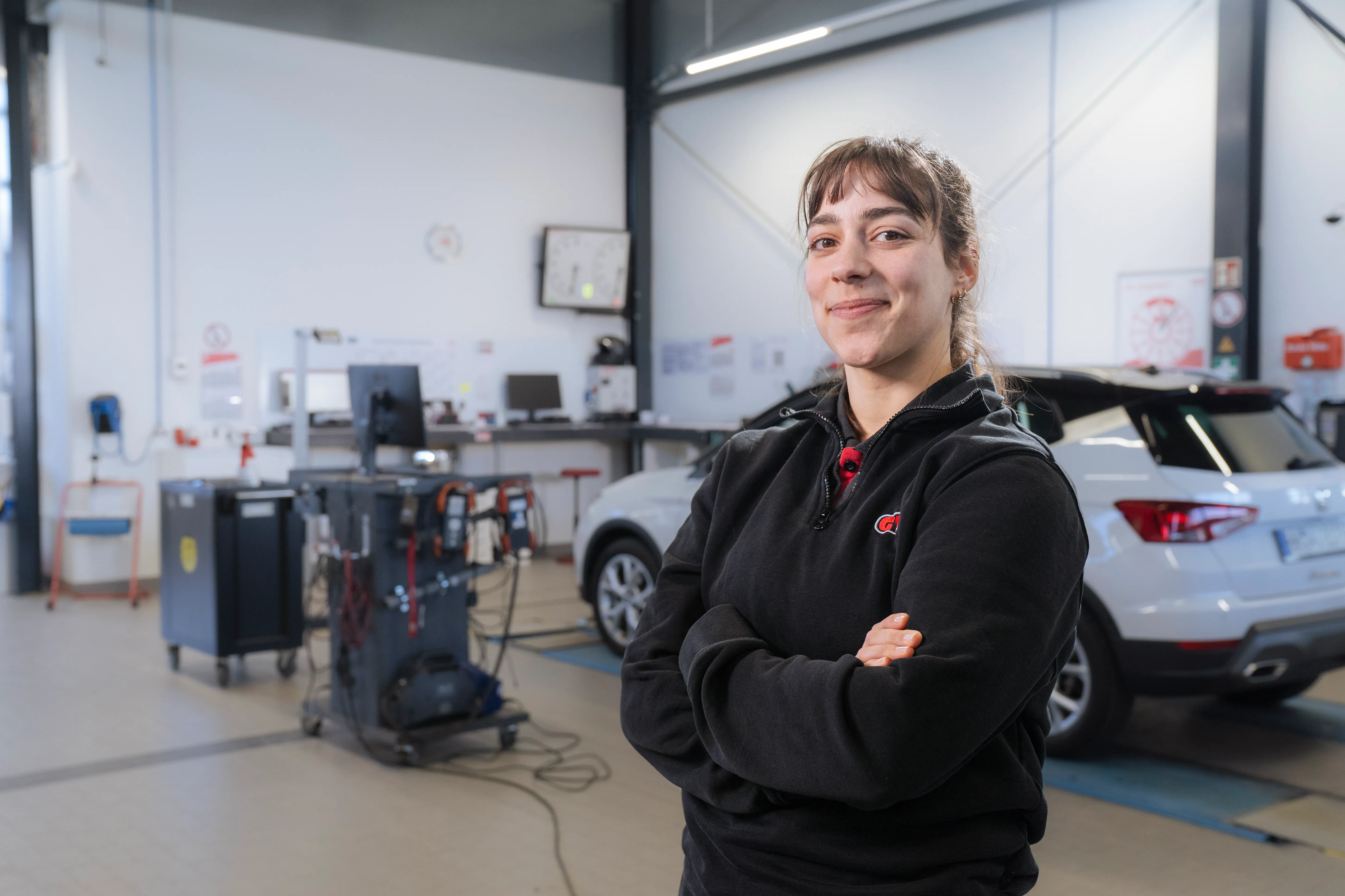 Ein lächelnder Mann in GTÜ-Uniform steht in einer Werkstatt und hält ein Diagnosegerät, im Hintergrund ein Auto auf einer Hebebühne.