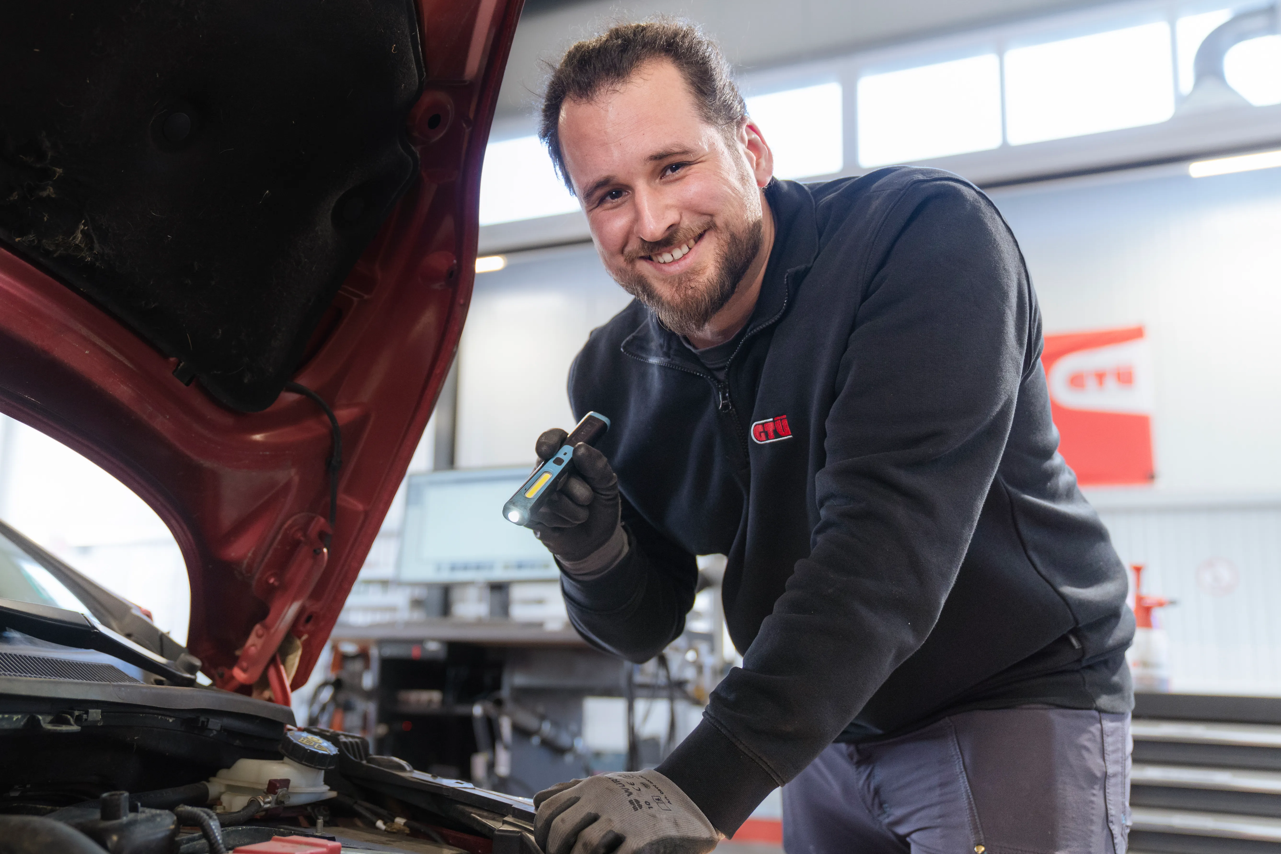 Eine GTÜ-Mitarbeiterin inspiziert ein Auto in der Werkstatt, während sie lächelt und Notizen macht.