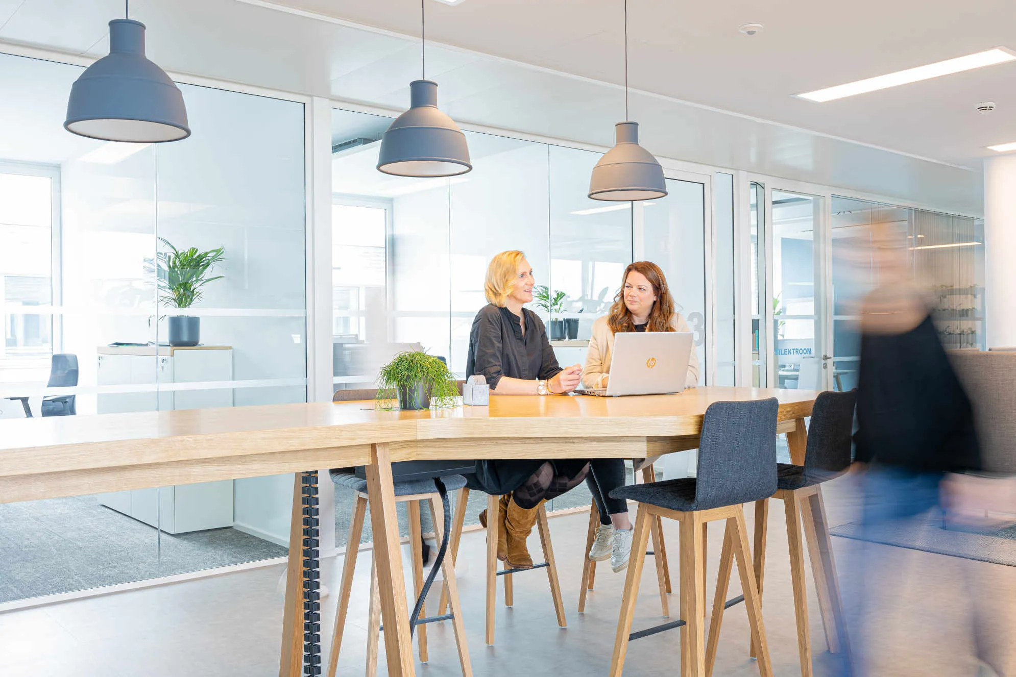 Zwei Frauen im modernen Büro tauschen sich am Tisch aus, Laptop vor sich – willkommen bei GWQ ServicePlus AG.