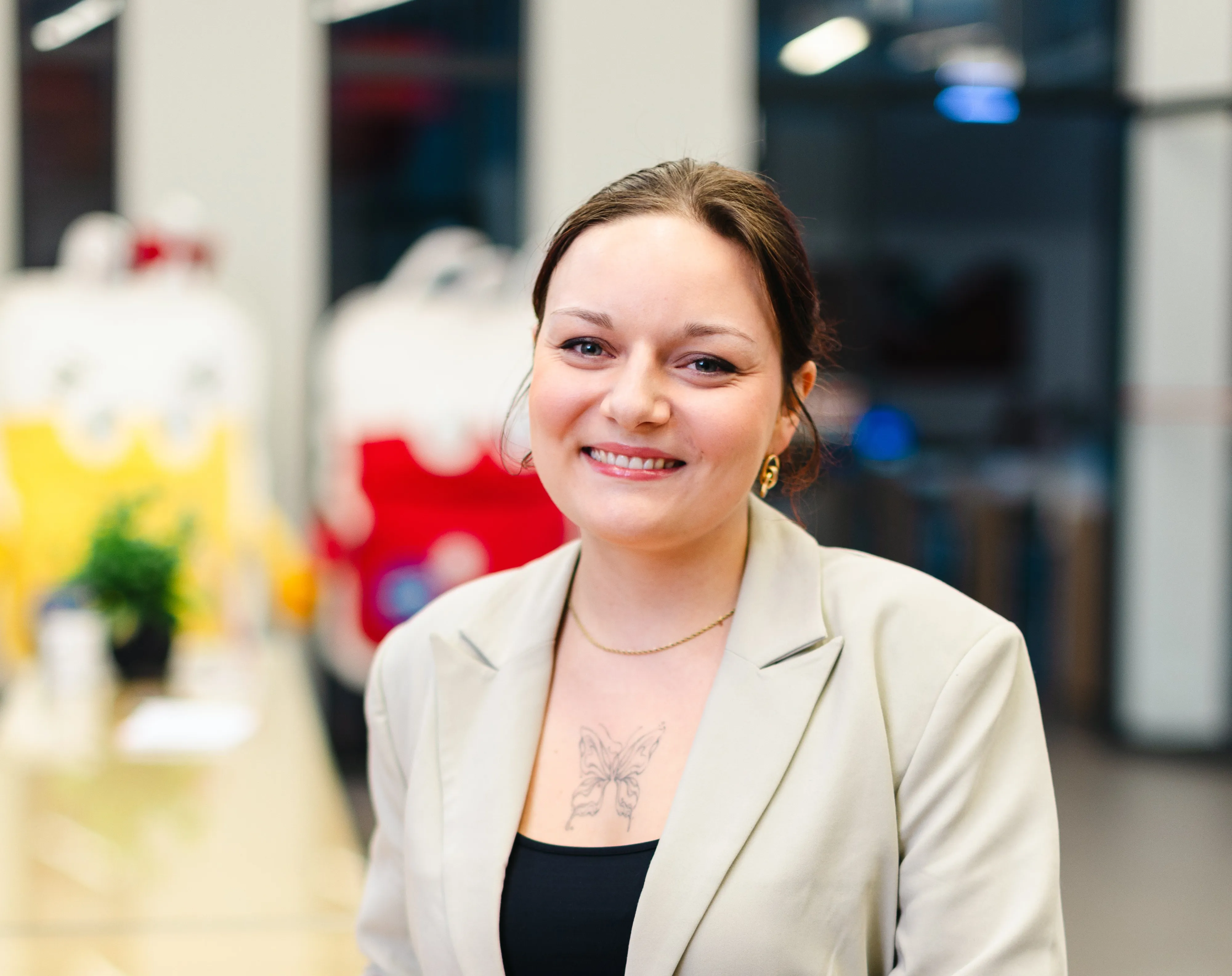Eine lächelnde Berufsstarterin von Haema steht im Foyer der Firmenzentrale. Hinter ihr verschwommen die Maskottchen des Blut- und Plasmaspendediensts