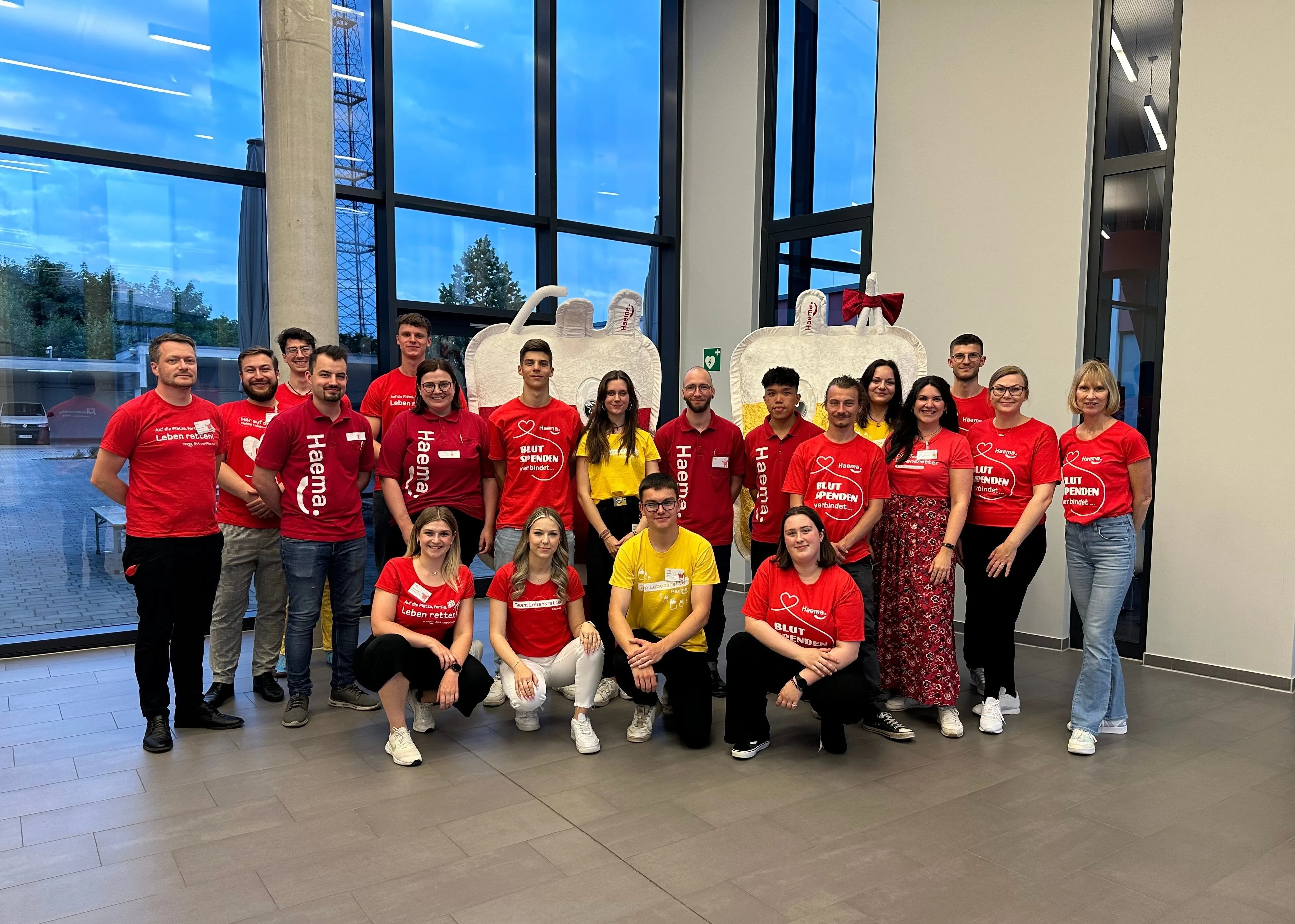 Gruppenfoto von Haema AG-Mitarbeitern in roten und gelben T-Shirts vor großen Blutspenden-Dekorationen, die freundlich in die Kamera lächeln.