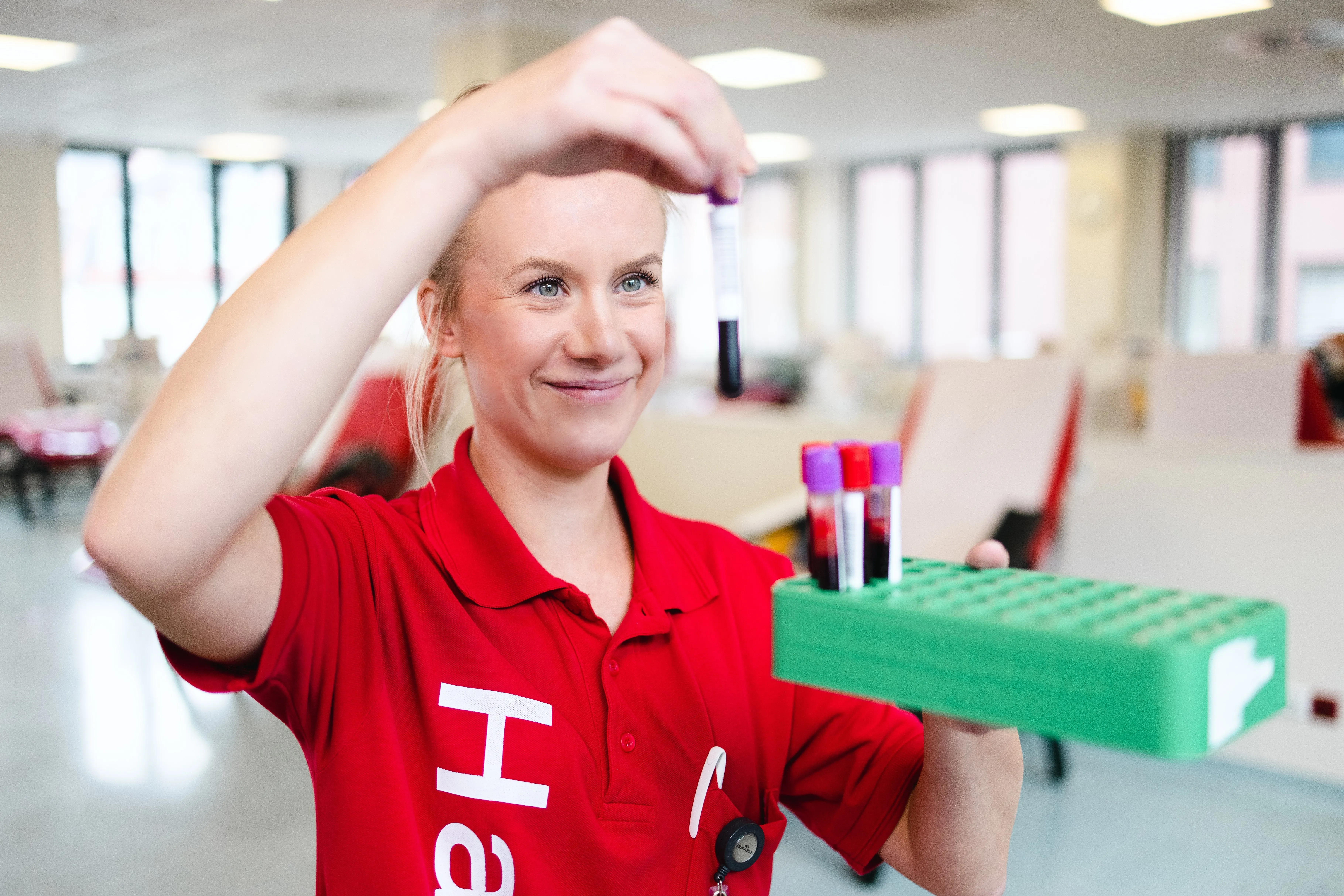 Eine Haema-Mitarbeiterin in roter Arbeitskleidung hält eine Blutprobe im Spendezentrum. Ein Haema Arzt berät einen Spender in einem Büro.