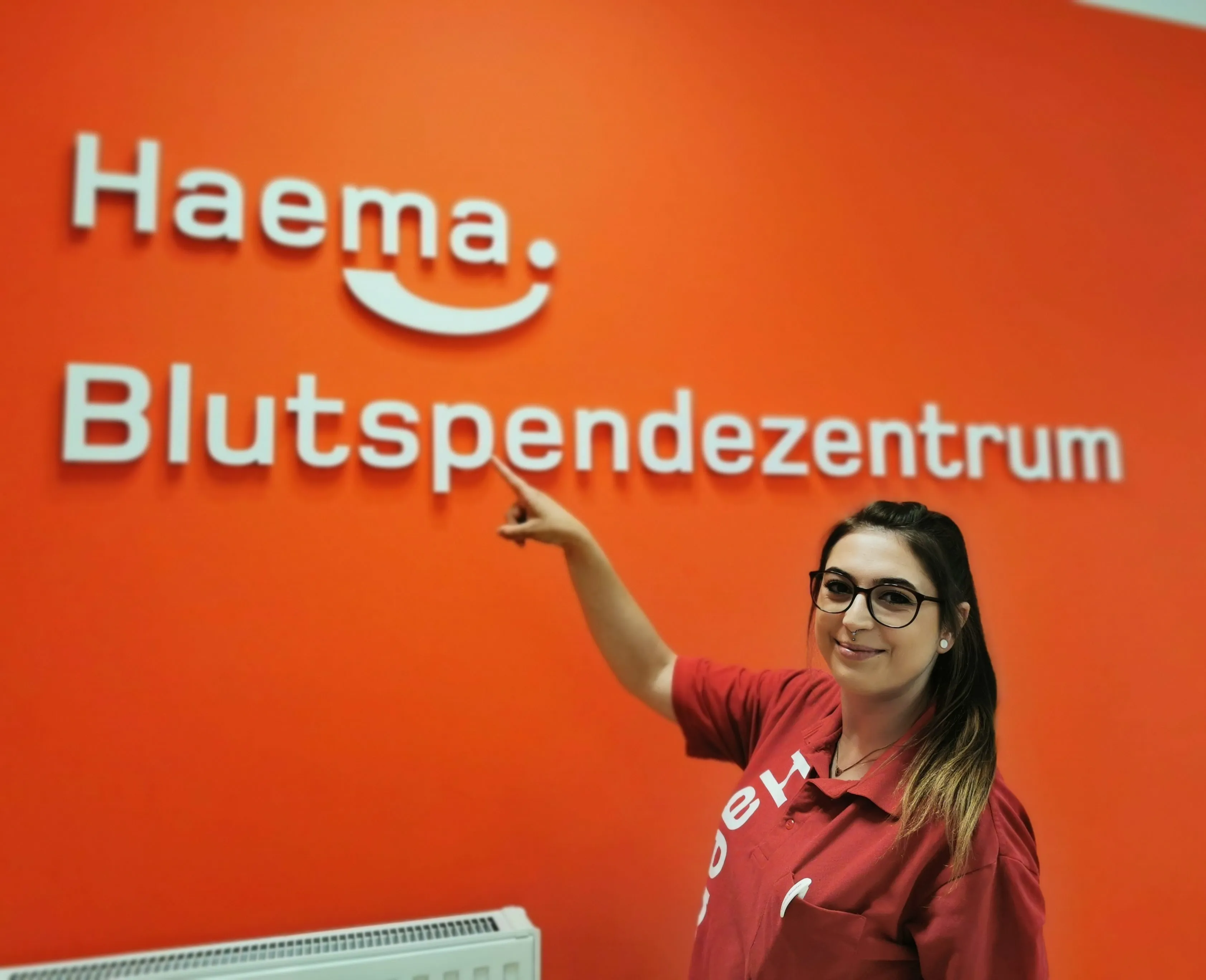 Haema Mitarbeiterin zeigt lächelnd auf rote Wand mit weißem Logo vor Haema Blutspendezentrum