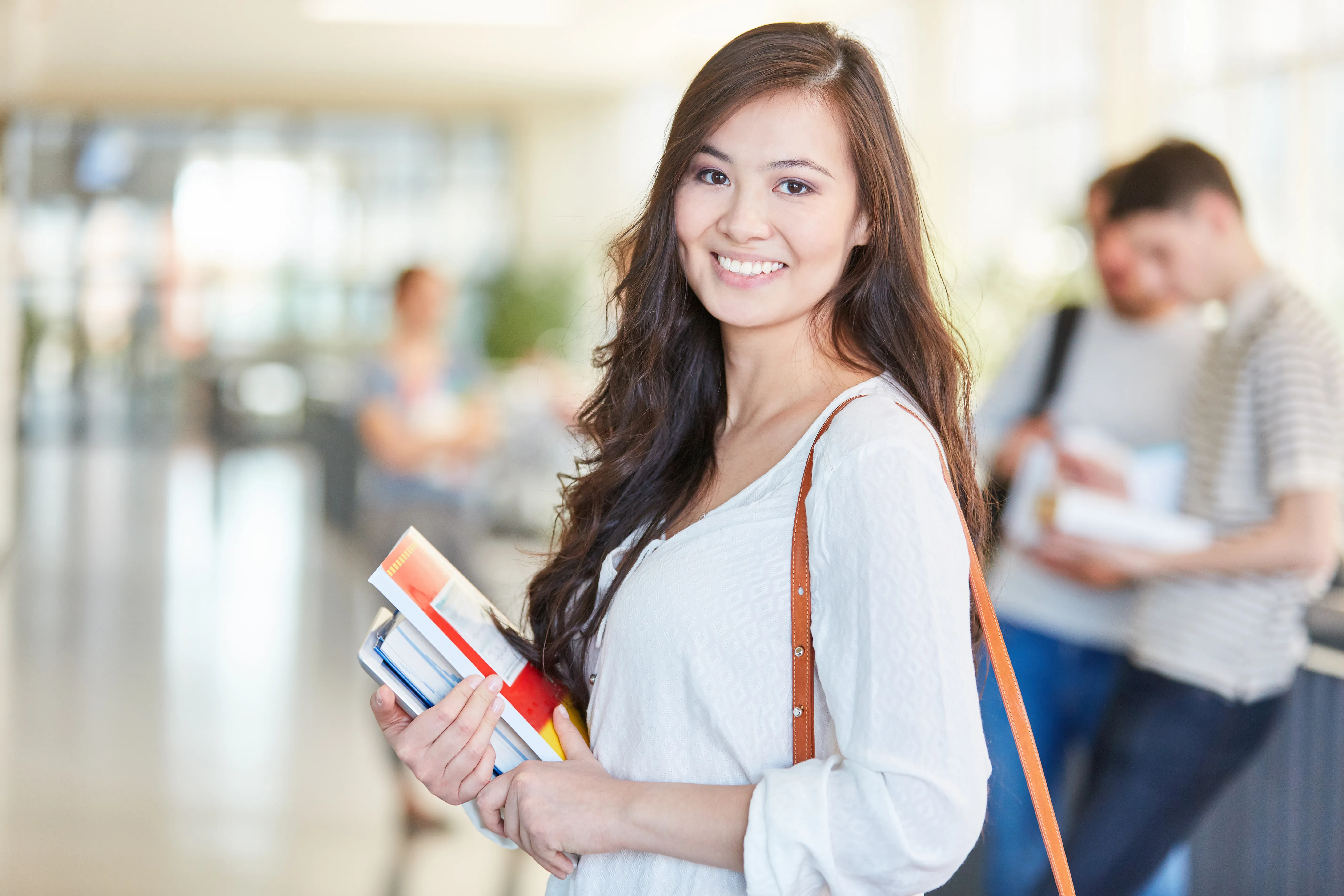 A woman smiling and holding books in a bright, modern hallway, embodying a positive and welcoming professional environment.