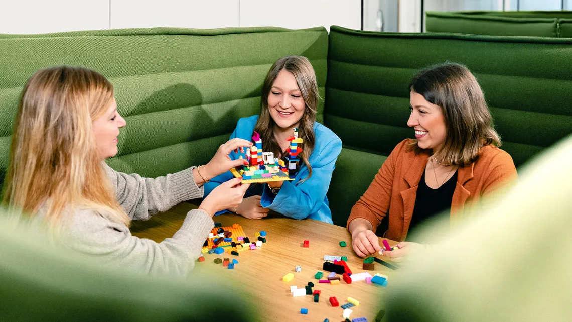 Drei Frauen bauen gemeinsam mit bunten Bausteinen an einem Tisch und lachen dabei.