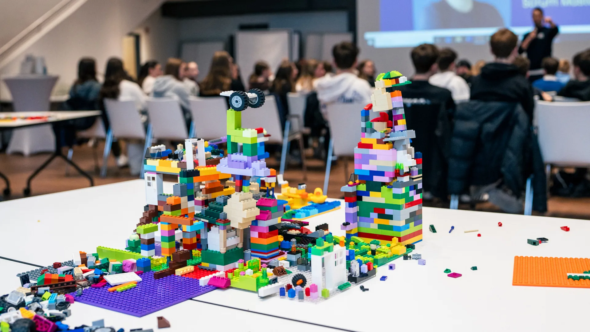 Lego creations on a table in a workshop, with a group gathered in the background listening to a speaker.