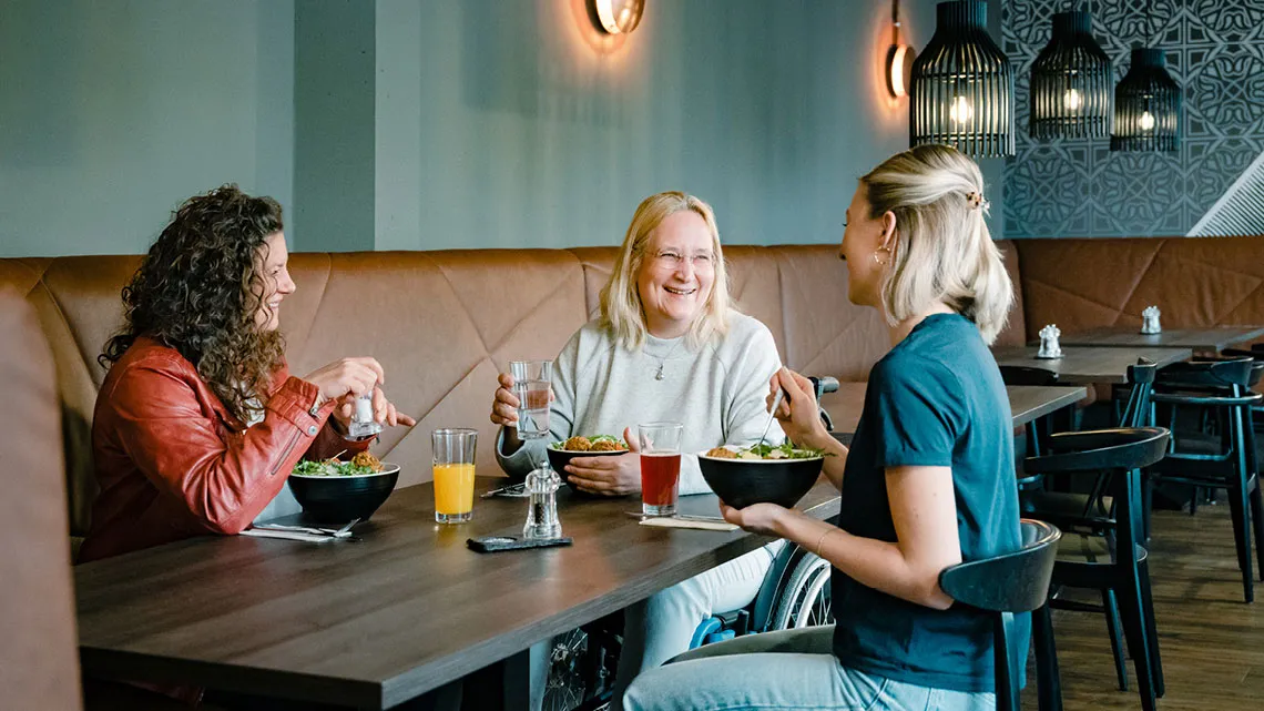 Drei Personen unterhalten sich in einem Café bei Getränken und Salaten. Eine Person sitzt im Rollstuhl.