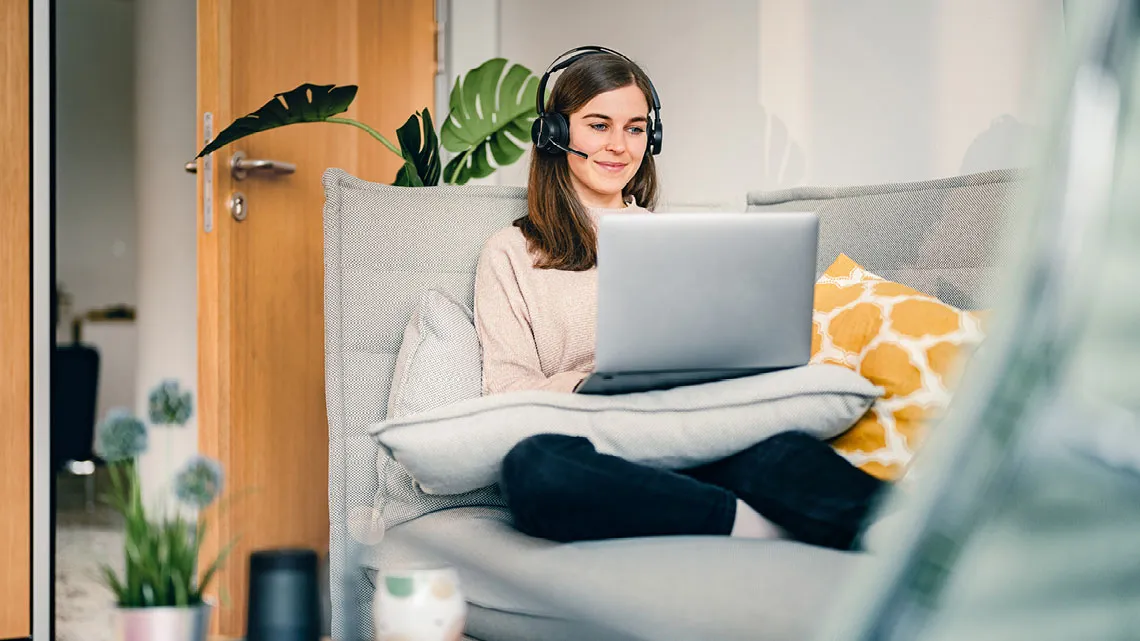 Eine Frau mit Headset arbeitet entspannt von zu Hause aus an einem Laptop, während sie auf einem Sofa sitzt.