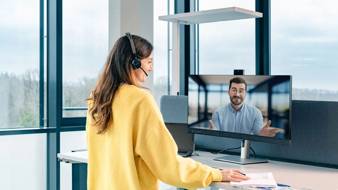 Eine Frau in einem gelben Pullover führt ein Videogespräch mit einem Kollegen auf einem Bildschirm in einem modernen Büro.