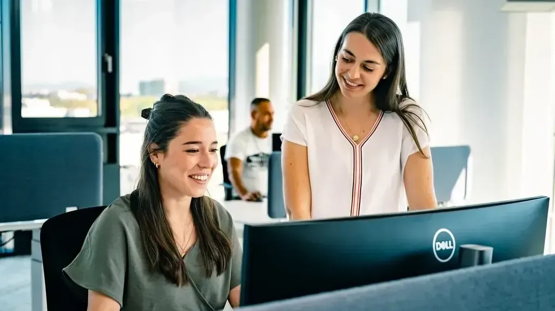 Zwei Frauen lächeln und arbeiten gemeinsam an einem Computer in einem modernen Büro bei Haufe-Group.