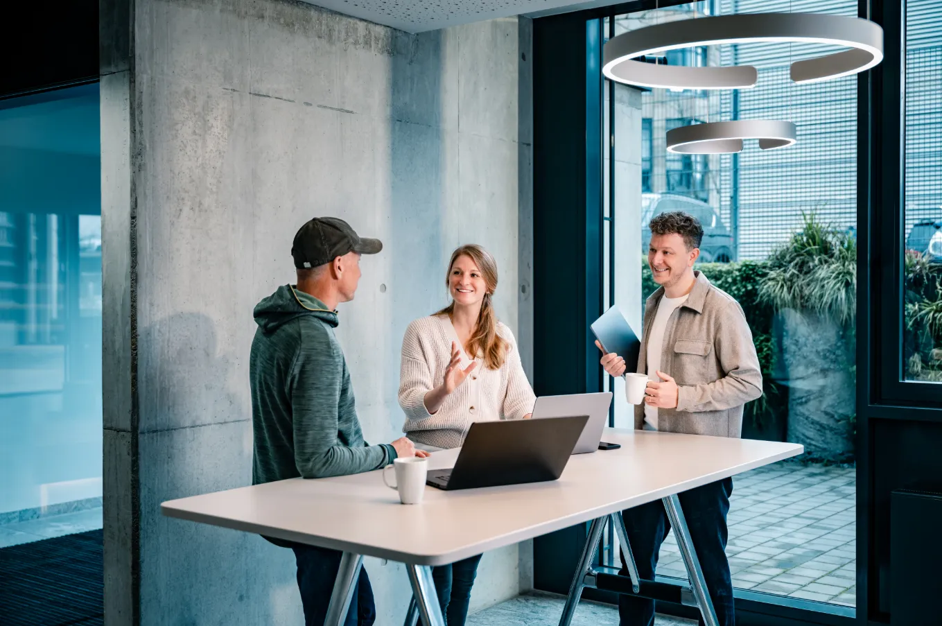 Drei Personen im Gespräch an einem Stehtisch in einem modernen Büro, mit Laptop und Tablet, lächelnd und entspannt.