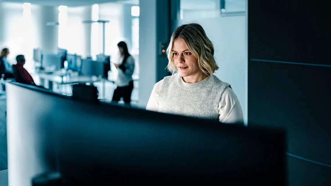 Eine Frau arbeitet konzentriert an einem Computer in einem modernen Büro, während Kollegen im Hintergrund beschäftigt sind.