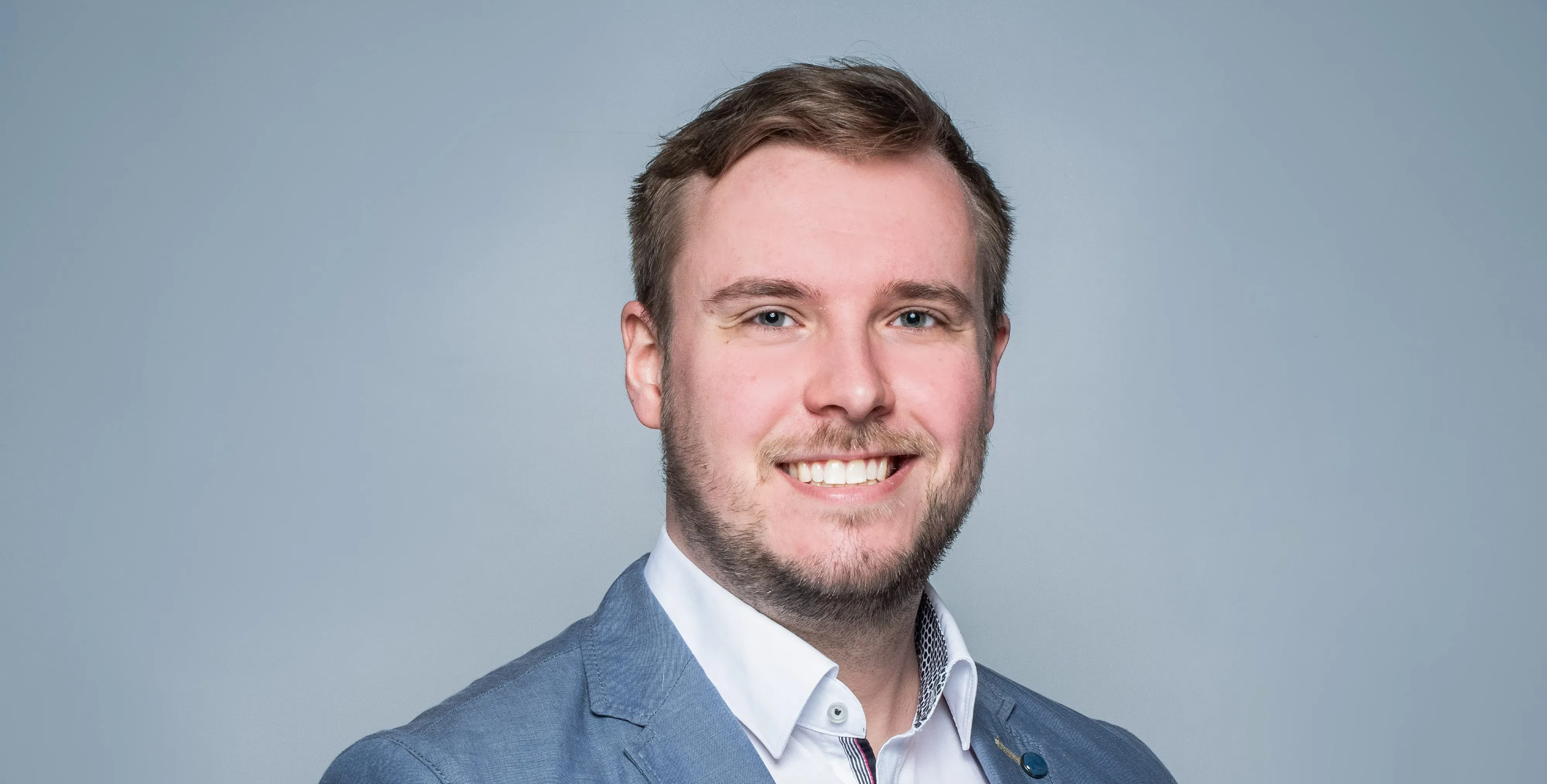 Zu sehen ist ein junger, freundlich lächelnder Mann in einem weißen Hemd und einem grauen Anzug