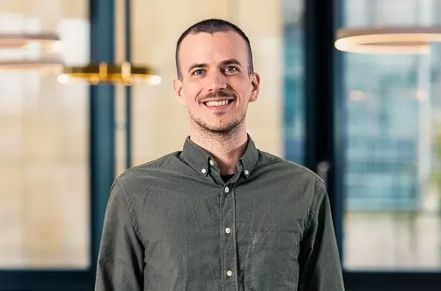 Zu sehen ist ein junger, freundlich lächelnder Mann in einem grauen Hemd.