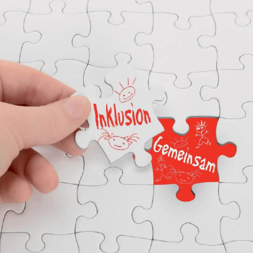 Eine Hand fügt ein Puzzlestück mit der Aufschrift "Inklusion" in ein Puzzle ein. Daneben ein rotes Stück mit "Gemeinsam".
