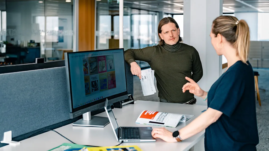 Zwei Kolleg:innen tauschen sich an einem modernen Arbeitsplatz über Medienprojekte aus, mit Magazinen und Computer.