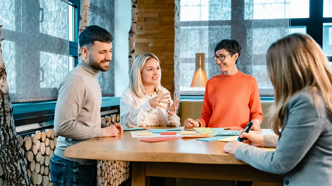 Vier Personen diskutieren an einem Tisch in einem modernen Büro, umgeben von bunten Notizen.