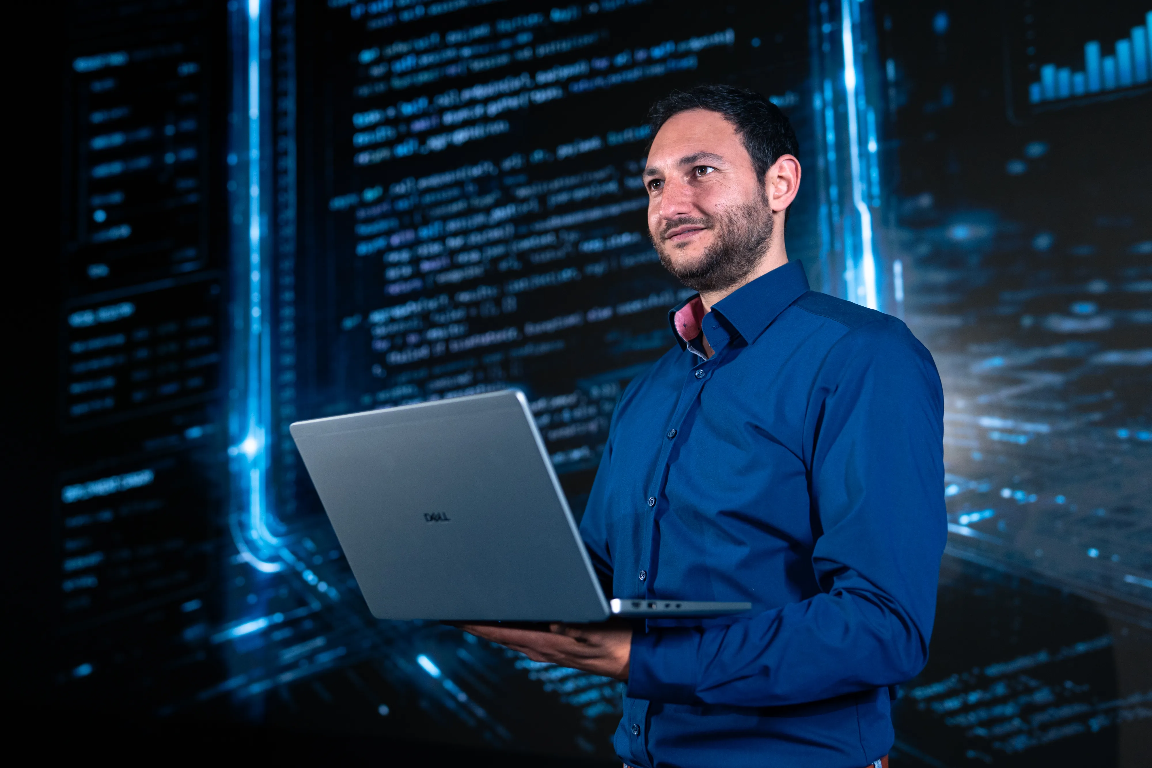 A smiling guy holding a laptop, standing in front of glowing digital code