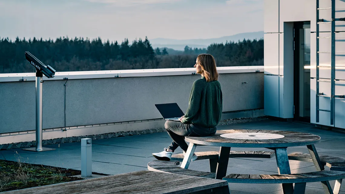 Eine Frau sitzt mit einem Laptop auf einer Dachterrasse und genießt die Aussicht auf einen Wald, ideal für kreative Pausen.