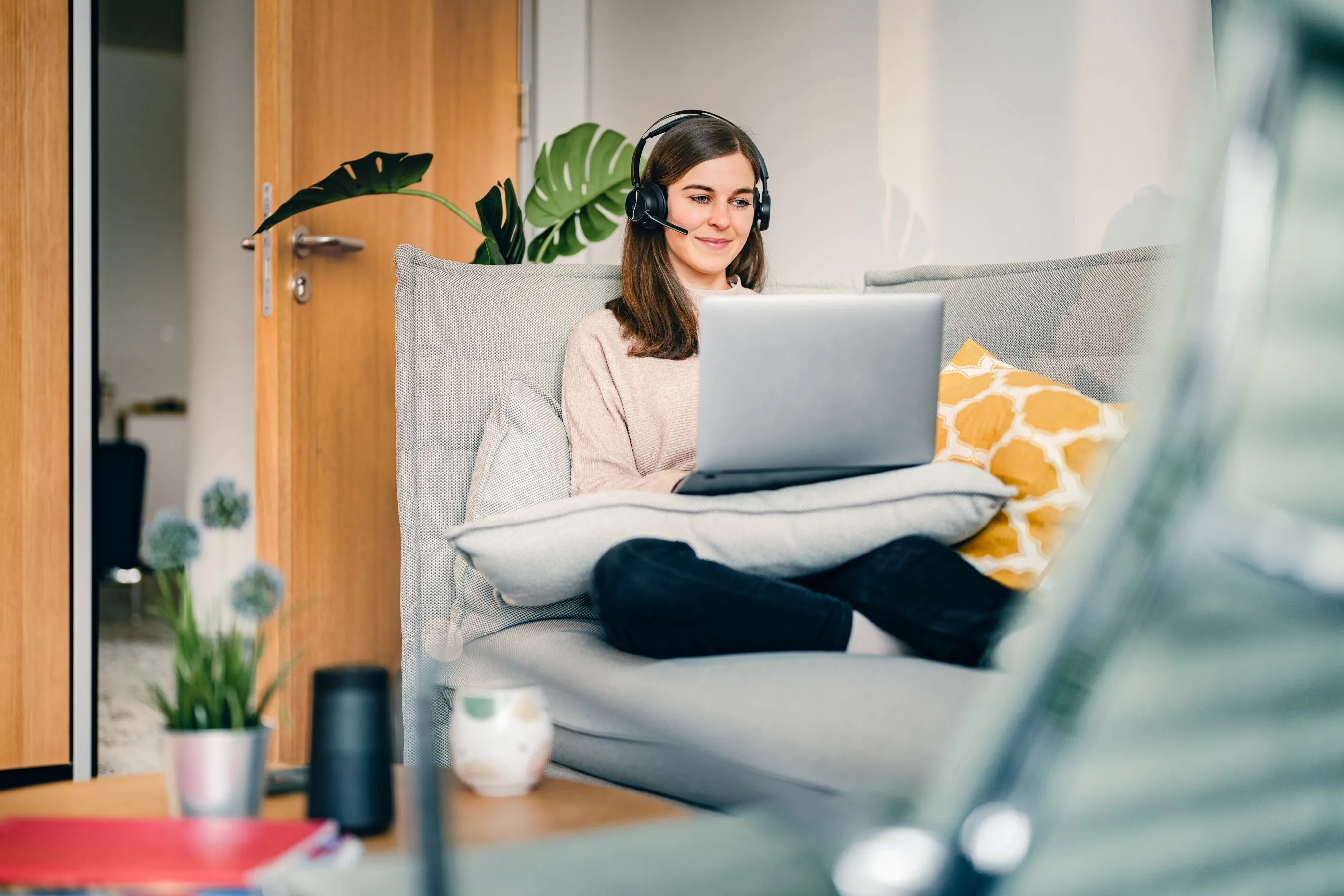Eine Frau arbeitet entspannt von zu Hause aus auf einem Sofa mit Laptop und Headset.