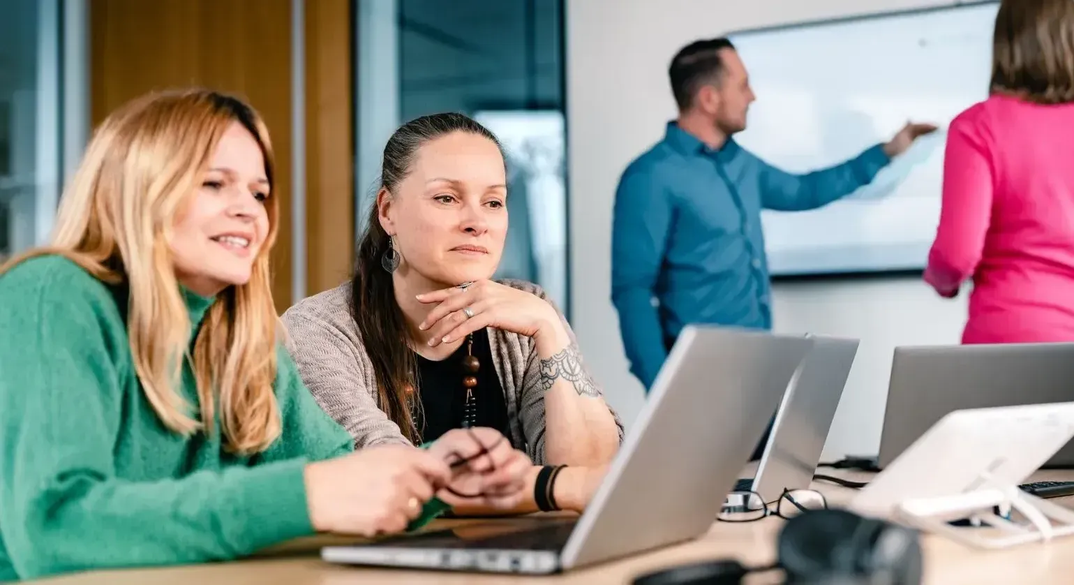 Zwei Frauen arbeiten am Laptop in einem Besprechungsraum, während im Hintergrund ein Kollege auf ein Whiteboard zeigt.