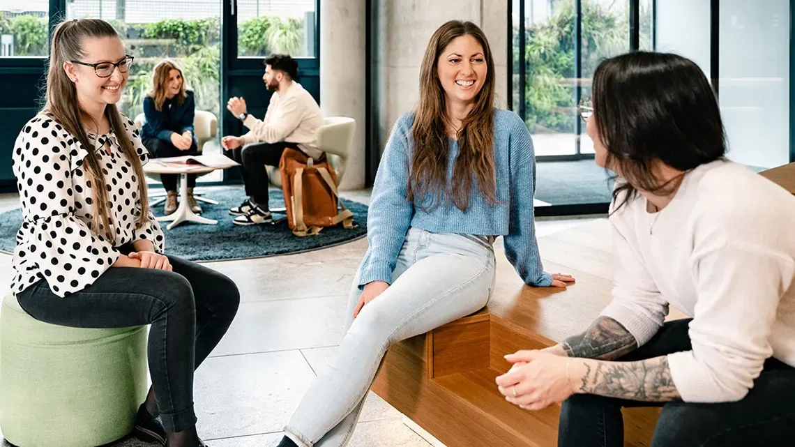 Colleagues in a modern office chat smilingly while another conversation takes place in the background.