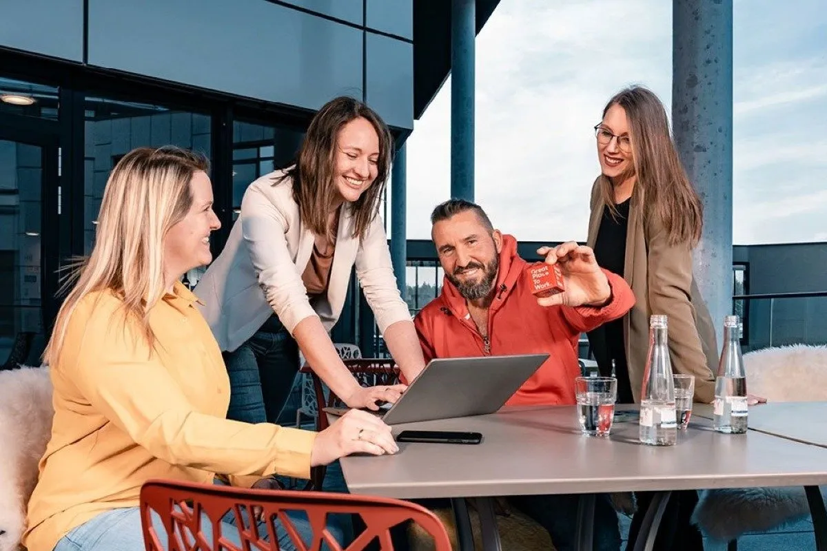 Vier Kollegen diskutieren fröhlich über ein Projekt an einem Laptop auf einer Terrasse.