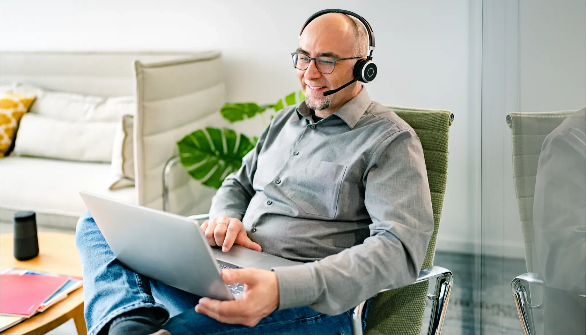 Ein Mann im Homeoffice, trägt ein Headset und arbeitet am Laptop. Entspannte Atmosphäre mit modernen Möbeln im Hintergrund.