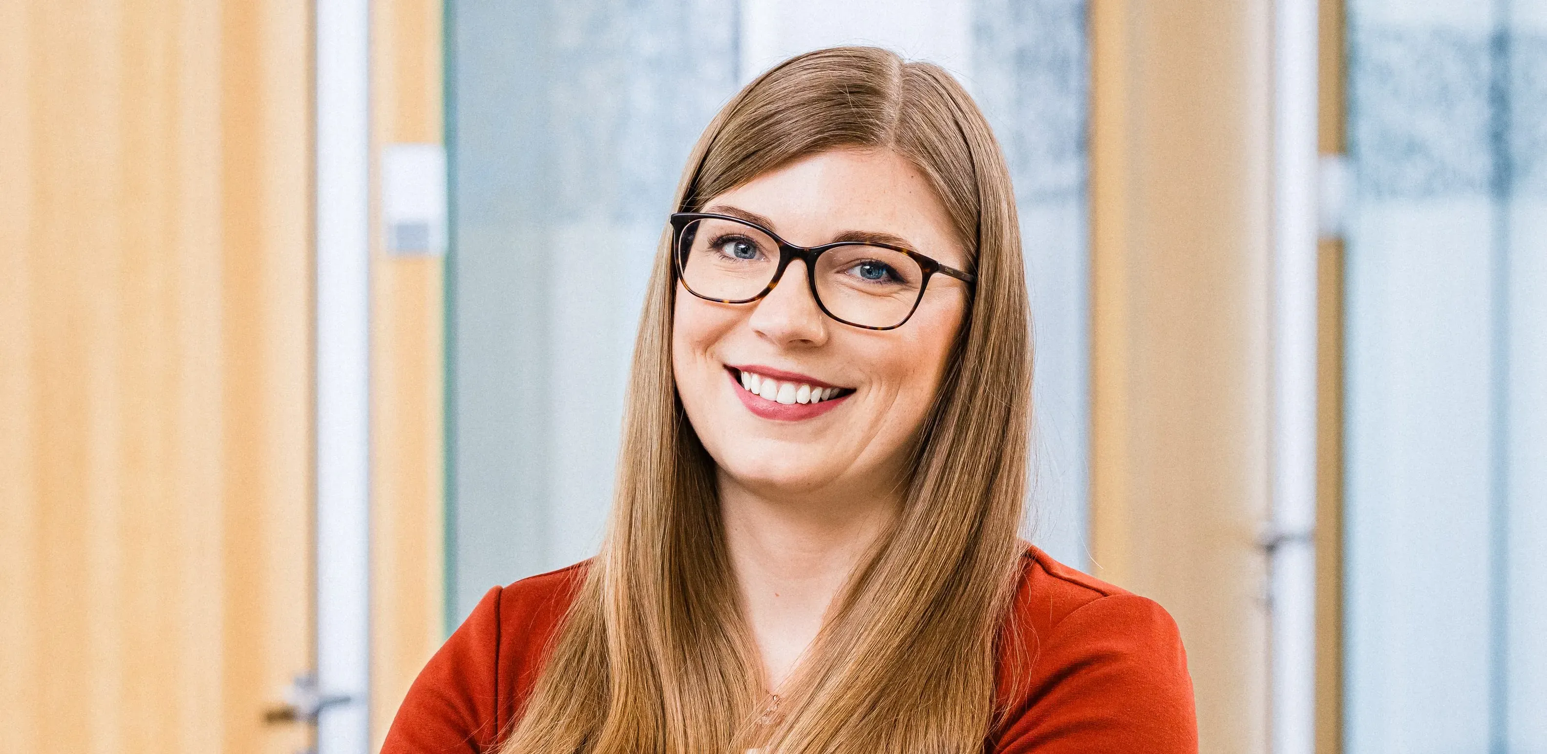 Eine lächelnde Frau mit Brille und rotem Blazer steht in einem modernen Büroflur.