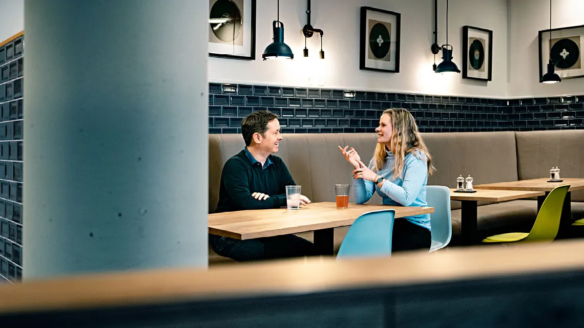 Ein Mann und eine Frau unterhalten sich lächelnd in einer modernen Cafeteria, beide haben ein Getränk vor sich auf dem Tisch.
