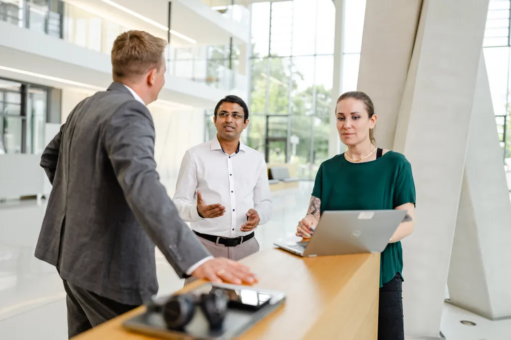 Ein Mann im Anzug spricht mit zwei Kollegen an einem modernen Stehpult. Eine Frau tippt auf einem Laptop.