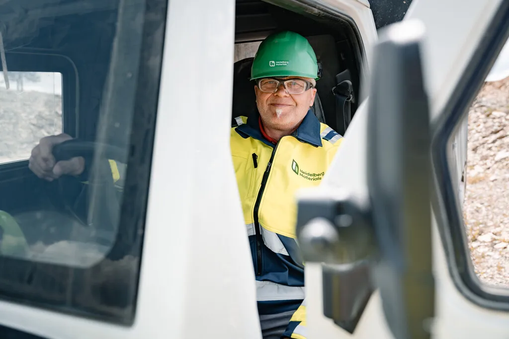 Ein LKW Fahrer mit Schutzhelm und Weste sitzt in einem Fahrzeug und lächelt.