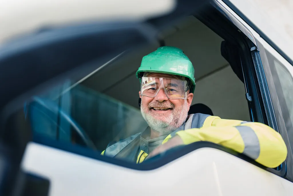 Ein LKW Fahrer mit grünem Schutzhelm und Schutzbrille lächelt aus einem Lkw-Fenster bei Heidelberg Materials GmbH.