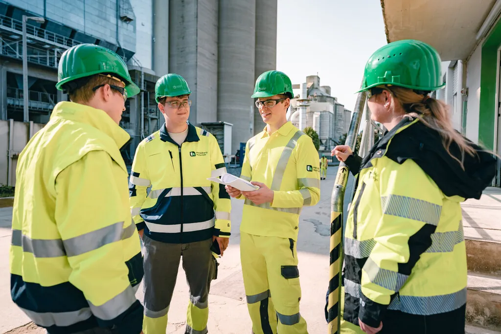 Vier Mitarbeiter in gelber Schutzkleidung und grünen Helmen besprechen Pläne auf einer Baustelle im Freien.