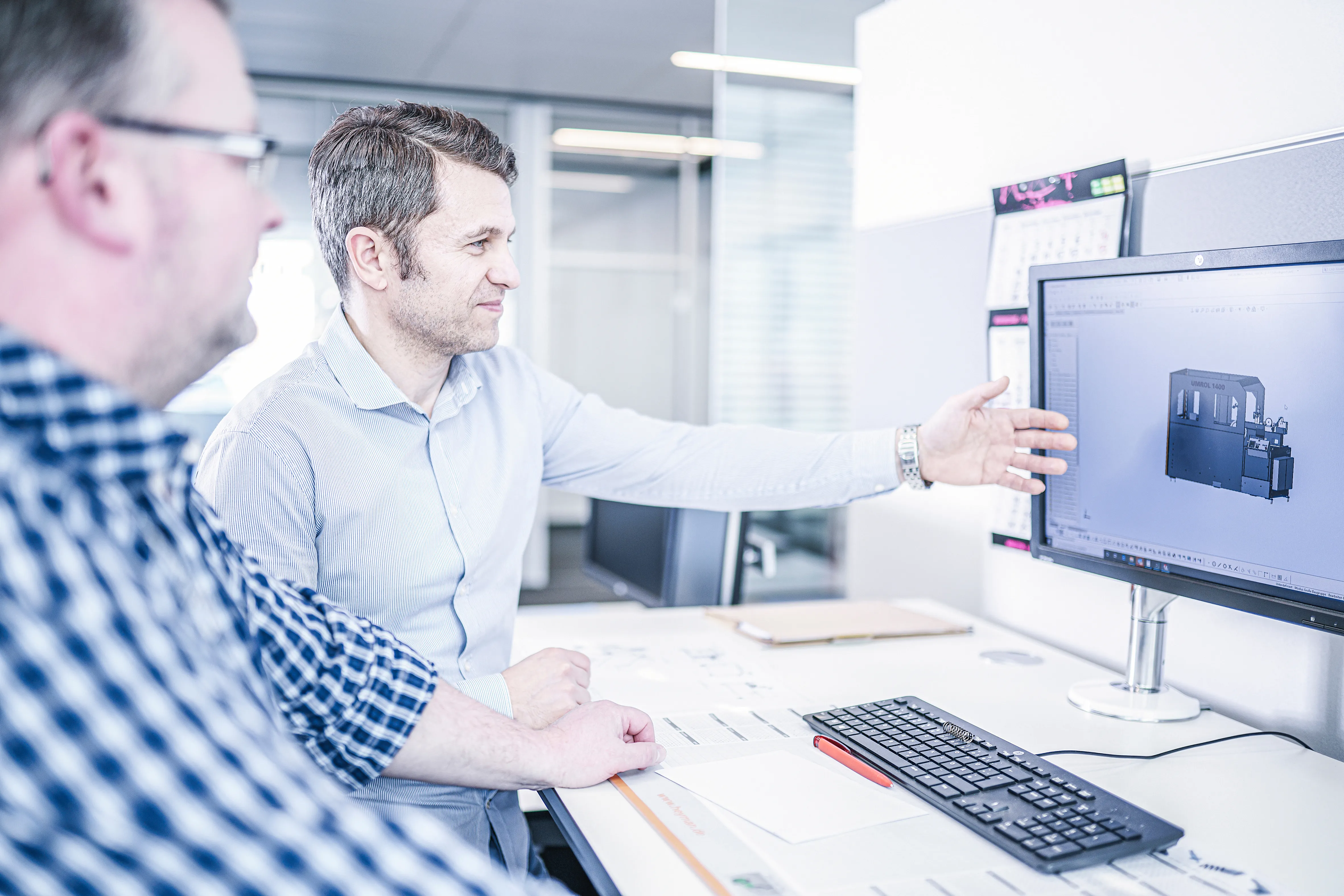 Zwei Männer in einem Büro diskutieren ein Maschinenmodell auf einem Computerbildschirm.