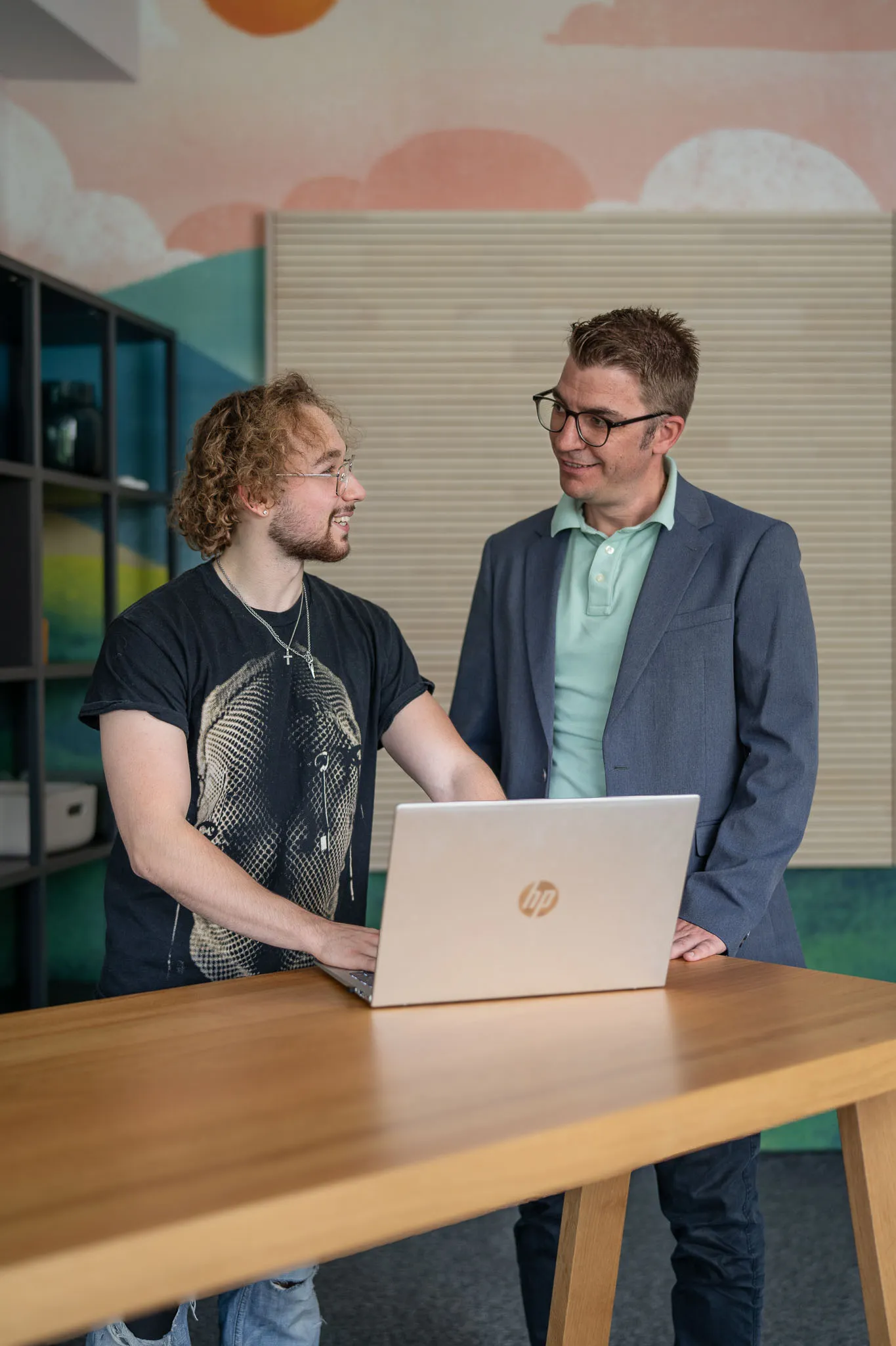 Zwei Kollegen bei Hochland unterhalten sich freundlich an einem Schreibtisch mit Laptop in modernem Büro.