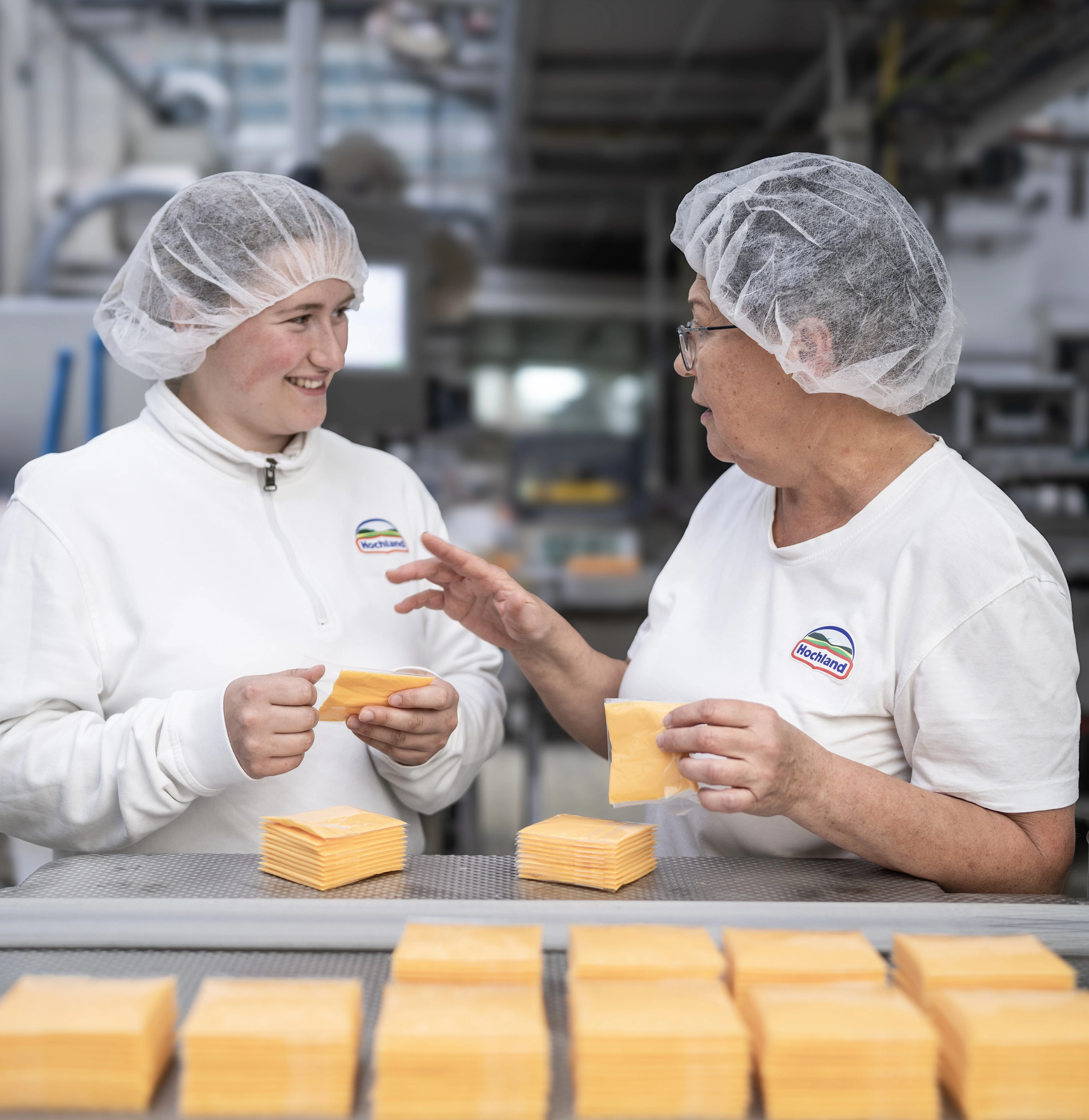 Zwei Hochland-Mitarbeiterinnen im Gespräch an einer Käse-Verpackungslinie, beide tragen Hygienekleidung und lächeln.