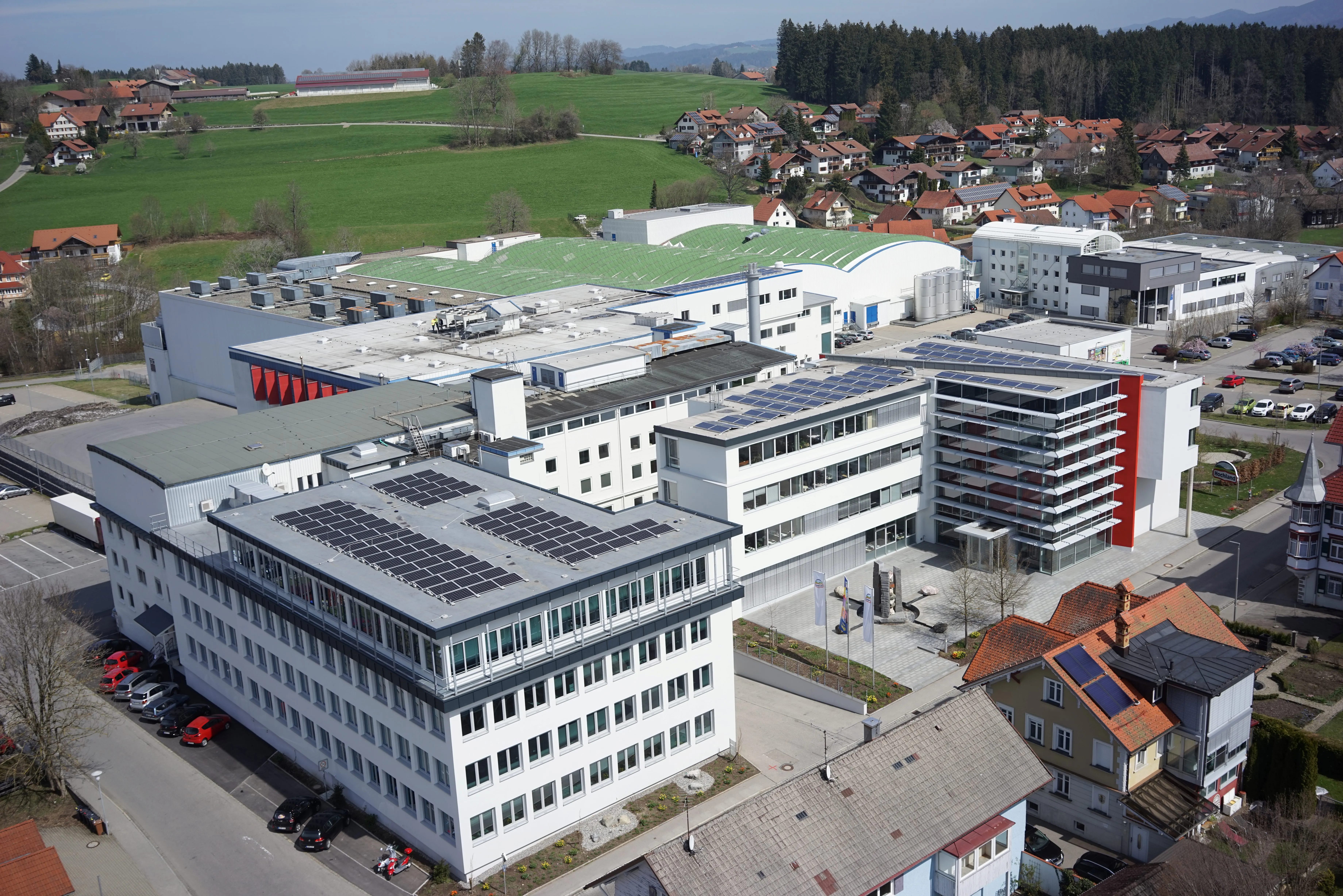 Moderne Hochland Firmenzentrale mit Solaranlagen auf dem Dach, umgeben von grüner Landschaft und Wohnhäusern.