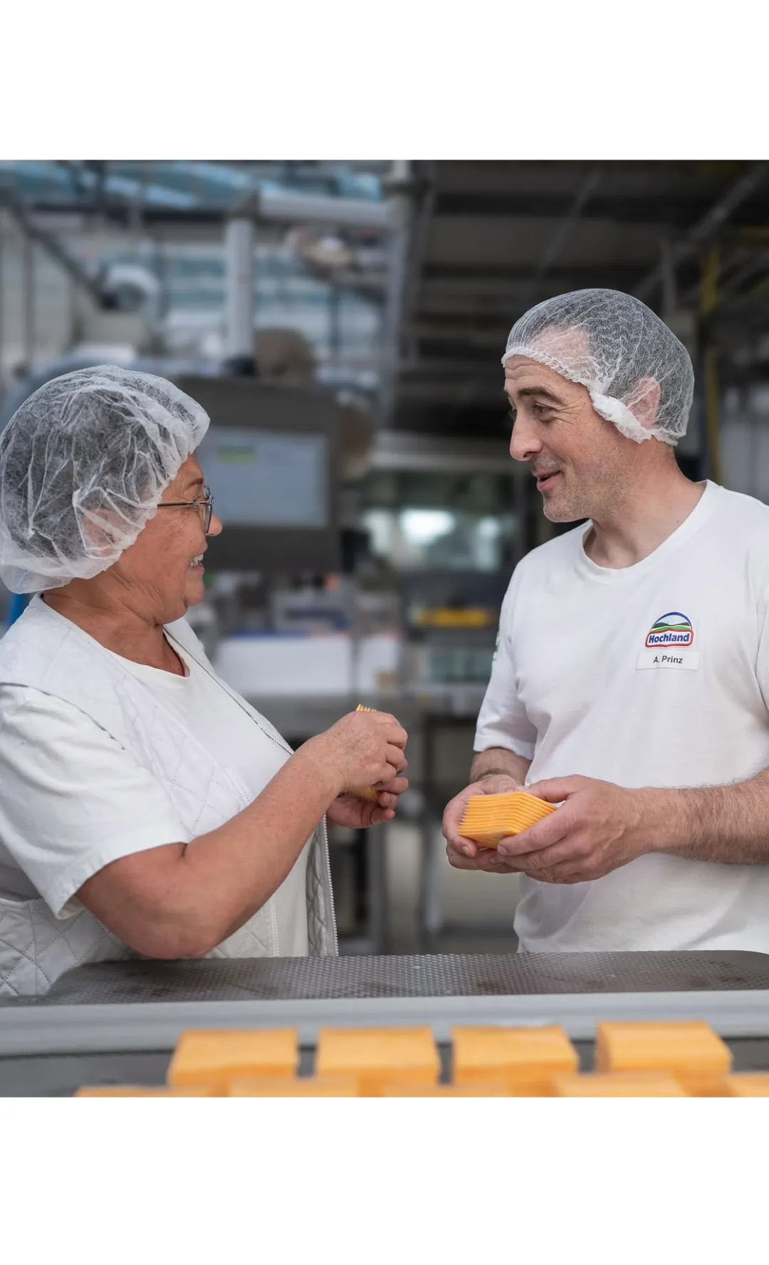 Zwei Mitarbeitende bei Hochland im Gespräch in der Produktion, beide tragen Hygienekleidung und halten Käsescheiben.