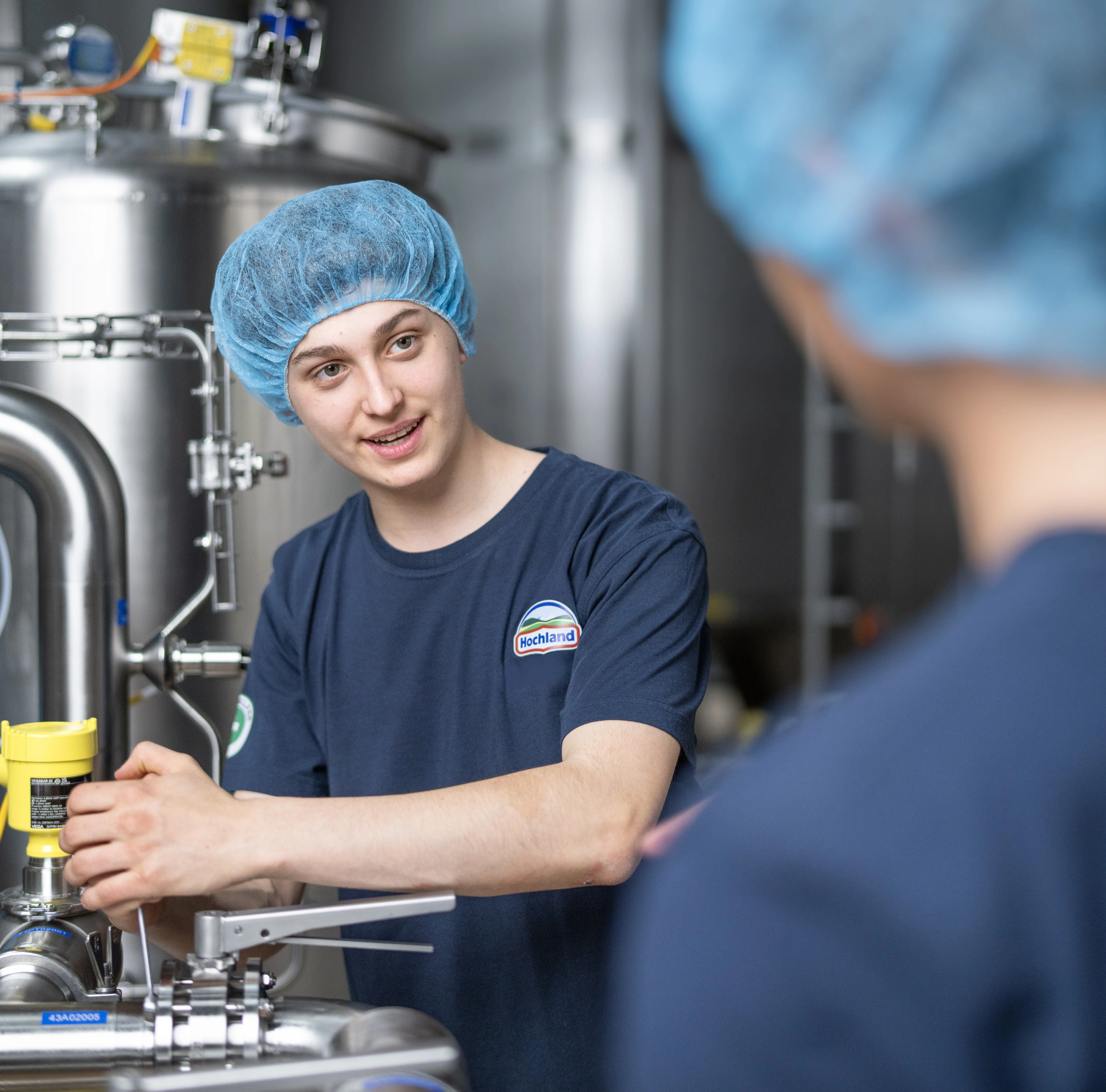 Junge Person im Hochland-T-Shirt arbeitet an einer Produktionsanlage und spricht mit einer Kollegin in der Lebensmittelindustrie.