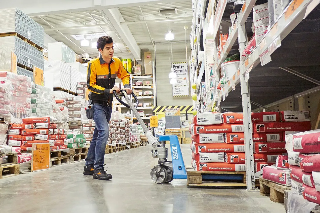 Mitarbeiter bei HORNBACH bewegt einen Stapelwagen in einem Baumarktgang, umgeben von Baustoffen und Regalen.