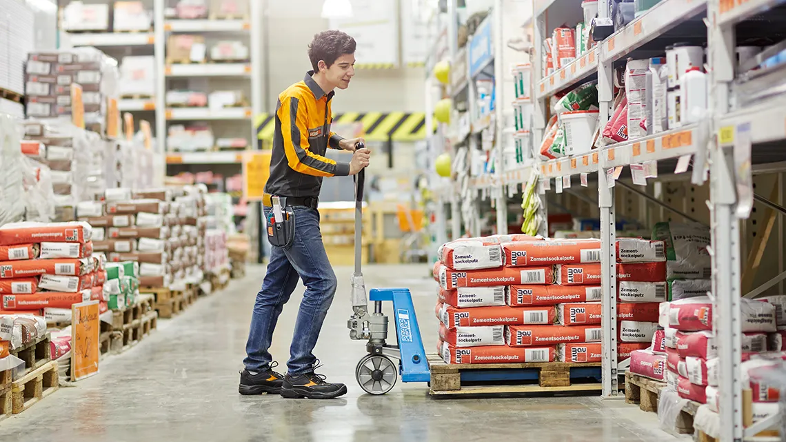 Un employé de HORNBACH déplace des sacs de ciment avec un transpalette dans un rayon de magasin, souriant et dynamique.